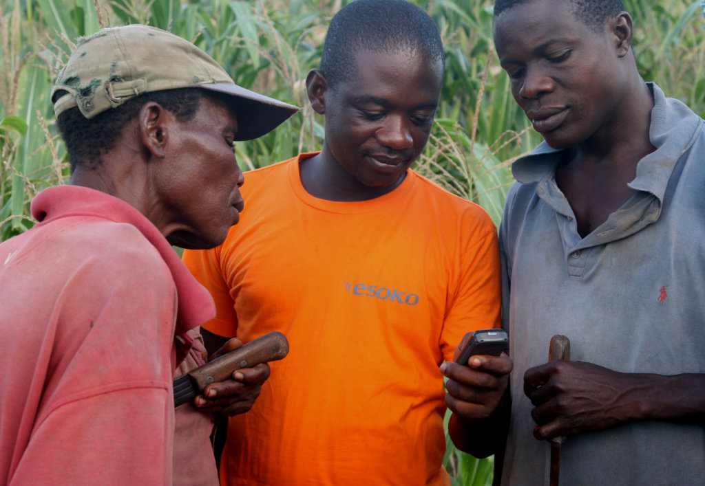 Three men stand together in a field with tall crops. The man in the center, wearing an orange shirt, is holding a mobile phone, which the others are looking at. Each man holds a tool, suggesting involvement in farming activities.