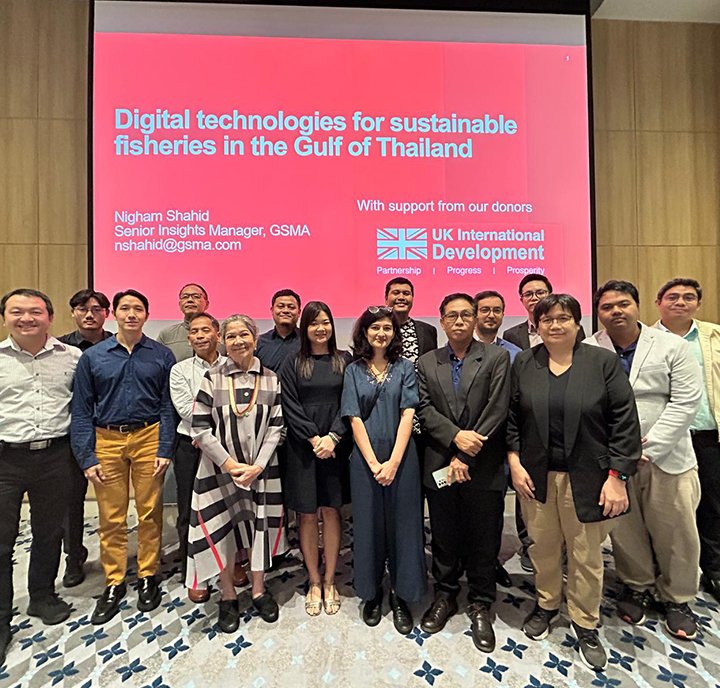 A group of people stands in front of a large screen displaying the text "Digital technologies for sustainable fisheries in the Gulf of Thailand" and logos of GSMA and UK International Development. They are in a conference setting.