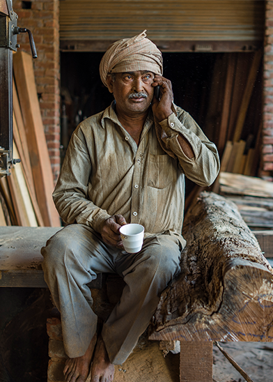 A man sits on a wooden beam in a workshop, talking on his cellphone. He wears a light brown shirt, gray pants, and a light-colored turban. He holds a white mug in his other hand. The background features wooden planks and a partially closed metal shutter.