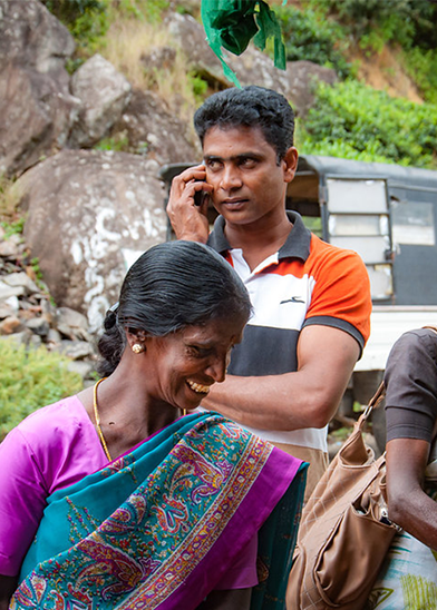 A woman with dark skin, wearing a vibrant sari, smiles broadly while standing outdoors. Behind her, a man in a red, white, and black shirt talks on a cellphone. The background features rocky terrain and greenery. A vehicle is partially visible to the side.