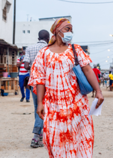 A woman wearing a colorful red and white dress and a headscarf walks on a sandy street. She is wearing a blue face mask and carrying a blue bag and some papers. Other people and buildings are visible in the background, with the street appearing busy and lively.