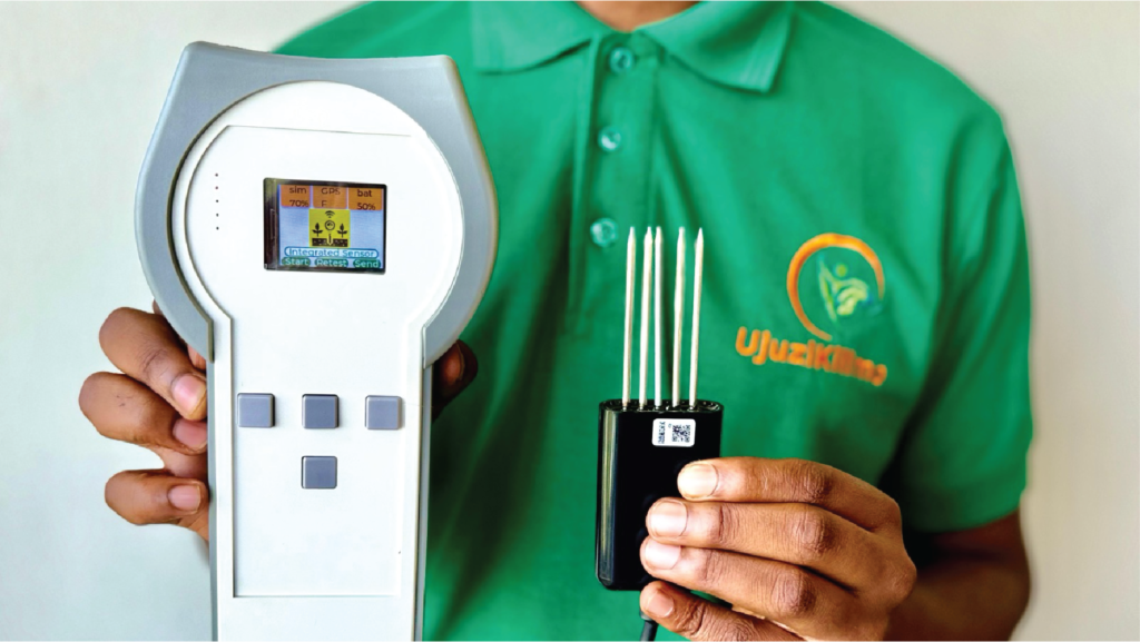 A person wearing a green shirt with a logo is holding two devices. The left device is a grey handheld monitor with a small screen displaying data. The right is a black rectangular sensor with four metal prongs. The shirt has an orange name and a leaf logo on it.