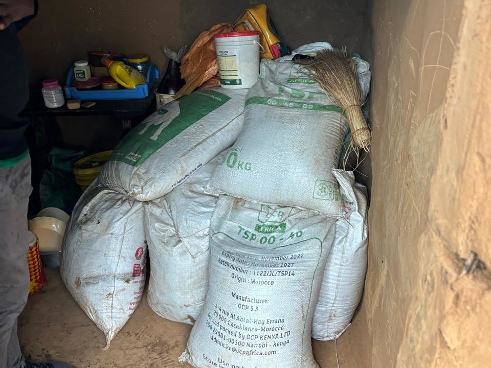 A small, dimly lit storage room with several large white sacks stacked on the floor against the wall. The sacks are labeled with text and appear to contain grains or produce. A few containers and jars are visible on a blue shelf nearby. A person stands partially visible on the left side.