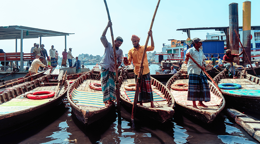 Three men stand on traditional wooden boats, holding long oars, with lively expressions. They wear patterned shirts and lungis. More people and boats are visible in the background, along with structures and the sparkling water under a clear sky. The scene is bustling and vibrant.
