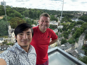 Two people taking a selfie in front of a glass barrier overlooking a lush, green landscape with trees and buildings. The person on the left wears a patterned shirt, and the person on the right wears a red Adidas shirt. The background features a winding road and a cloudy sky.
