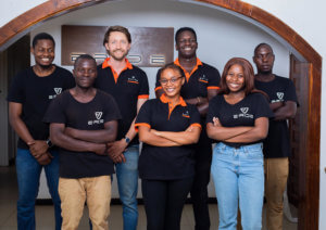 A group of seven people stands smiling indoors beneath a wooden arch. They are wearing black shirts with a logo that says "E-RIDE" in white and orange. The group includes men and women of diverse backgrounds, with some crossing their arms playfully. They appear relaxed and friendly.