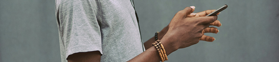 A person in a grey t-shirt holds a smartphone, earphones visible. Two wooden beaded bracelets adorn their left wrist against a plain grey background. The focus is on their hands and the phone, capturing a moment of tech interaction and computation at its simplest.