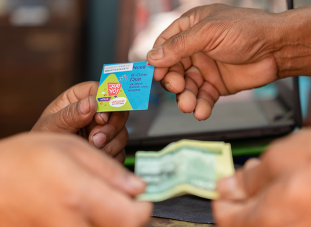 Close-up of hands exchanging a small, colorful packet with Nepali script for paper currency. The packet has a blue and pink design, while the money is green. A blurred electronic device is in the background. The scene suggests a transaction or purchase.
