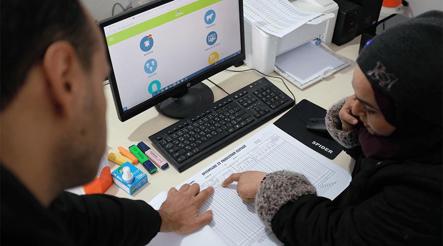 A person in a black jacket points at a document labeled "Restaurant Attendance Sheet" on a desk, reminiscent of bringing efficiency to livestock management: lessons from MooMe. Nearby, someone in a headscarf and dark coat looks on. A computer screen showing a colorful dashboard and office supplies are visible in the background.