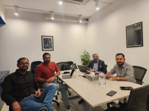Four men are seated around a light wooden table in a modern, well-lit office. They have laptops and documents in front of them. The background features minimalist decor, including framed art and a potted plant. The atmosphere appears professional and collaborative.