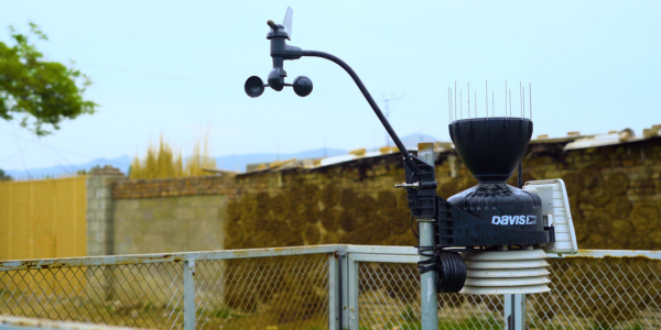 A black weather station with sensors and anemometer is mounted on a metal fence. The device has the label "DAVIS" and is surrounded by a yard with stone walls, fencing, trees, and distant mountains under a cloudy sky.