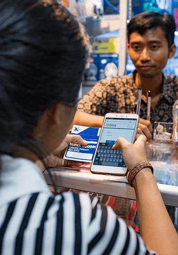 A person is sitting at a counter, holding a smartphone displaying a BNI banking app. The screen is in focus as they type. In the background, a vendor sits opposite, surrounded by phone accessories. The person has a striped shirt and the vendor wears a patterned shirt.