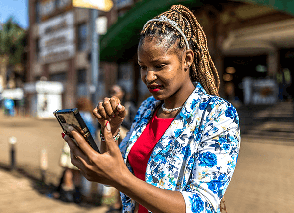 A woman with braided hair looks at her phone while standing outside. She wears a floral blazer over a red top and a pearl necklace. The background shows a blurred urban setting with buildings and a walkway. Her expression is focused.