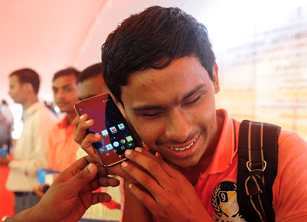 A young man wearing a pink shirt, holding a large smartphone to his ear, smiles slightly. Behind him, a few people stand in line, and a hand is pointing at the phone screen. The background is indoors with an orange tint and blurry text.