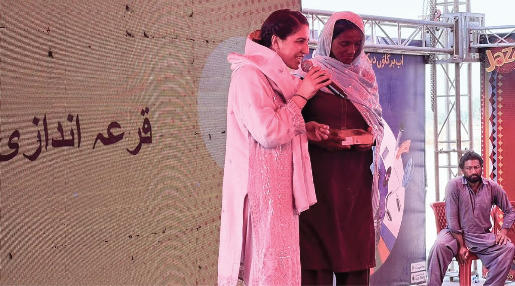 Two women stand on a stage, one speaking into a microphone, the other holding a box. They wear traditional South Asian attire. Behind them is a large screen displaying text in Urdu. A seated man observes from the side. The event appears to be outdoors with a metal frame structure.