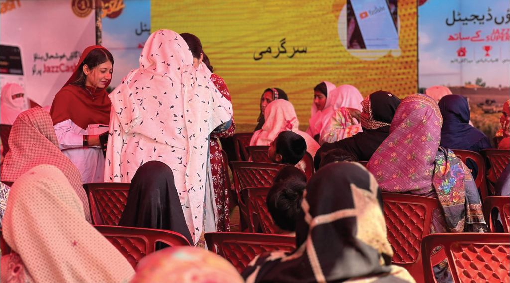 A group of women and children in vibrant traditional attire sit on red plastic chairs. They are gathered outdoors, with a large yellow screen in the background displaying Urdu text. Some women are engaged in conversation, while others face the screen. The setting suggests a community event.