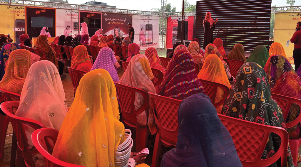 A large group of women wearing vibrant, multicolored headscarves sit on red plastic chairs facing a stage. A person speaks onstage near a black panel. The setting is outdoors, with posters and banners in the background, suggesting a community or cultural event.
