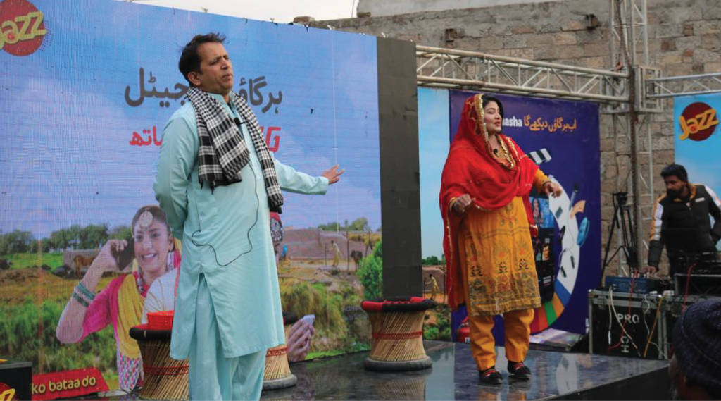 A man and a woman perform on an outdoor stage. The man wears a light blue traditional outfit and a black-and-white checkered scarf. The woman wears an orange and yellow traditional dress with a red scarf. A large screen behind them displays promotional content. People watch nearby.