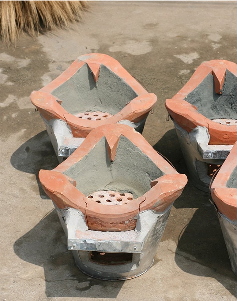 Four traditional clay and metal charcoal stoves are arranged on a concrete surface. Each stove has a reddish-brown clay top with a central hollow and circular air holes for ventilation. The bases are metallic, providing sturdy support for cooking activities.