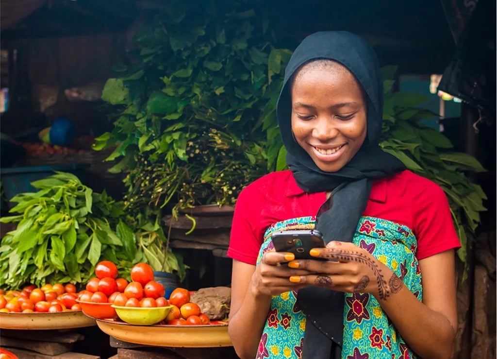 A smiling woman in a colorful dress and black headscarf looks at her smartphone. She stands in front of a market stall filled with tomatoes and leafy greens. The produce is displayed in bowls, creating a vibrant background of fresh vegetables.