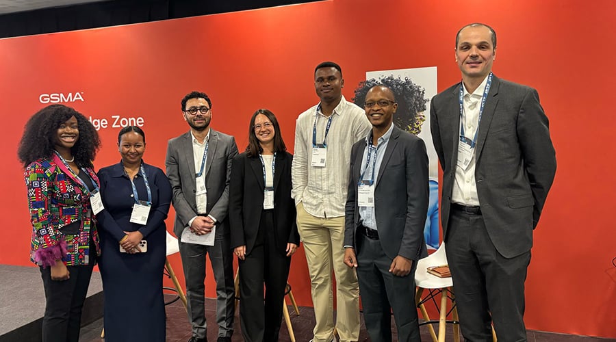 Seven people in business attire stand smiling against a red wall with "GSMA Knowledge Zone" visible. Sporting name tags, the group of four men and three women exudes a professional yet welcoming atmosphere. In the background, three white chairs indicate a space where deep tech insights are likely shared.
