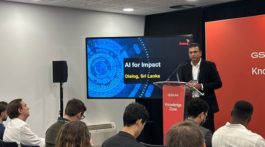 A man in a suit stands at a podium presenting to an audience. Behind him, the screen displays "AI for Impact, Dialog, Sri Lanka" with a blue digital graphic. Taking place in the GSMA Knowledge Zone, this deep tech event captivates attendees who are seated and listening attentively.