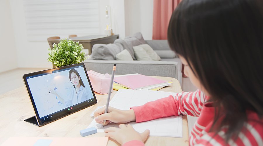 A girl in a pink striped shirt is sitting at a wooden desk, taking notes while watching an online class on a tablet. The screen shows a woman teaching. There are notebooks, a pink pencil case, and an eraser on the desk. A plant and a couch are visible in the background.