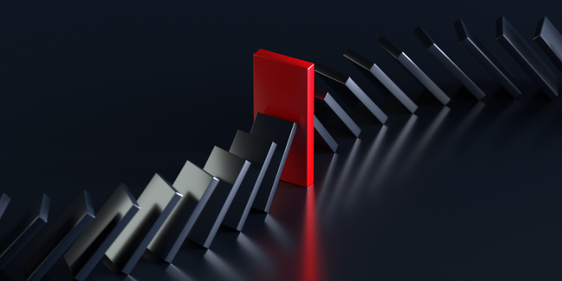 A row of black dominoes falling in a sequence from left to right on a glossy black surface, interrupted by a single standing red domino in the center, preventing the rest of the dominoes from toppling. The scene is set against a dark background, creating a dramatic contrast.