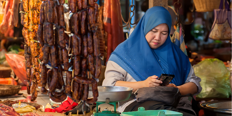 A woman wearing a blue hijab sits at a market stall, looking at her phone. In front of her are hanging dried sausages and a metal scale, with various market goods and bags visible in the background. The scene is colorful and bustling.