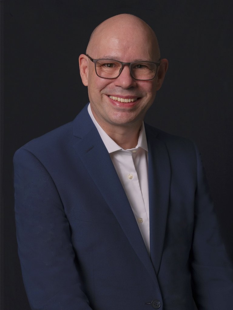 A smiling bald man wearing glasses, a white shirt, and a navy blue suit jacket poses against a plain dark background. He is facing the camera with his hands down, exuding a friendly and professional demeanor.