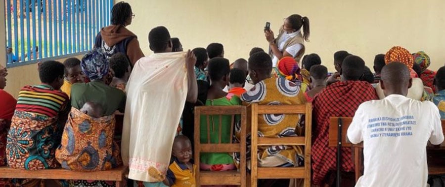 A group of adults and children, mostly women in colorful clothing, sit on wooden benches facing a woman standing and taking a photo with her phone. The setting appears to be indoors, with blue bars on the window to the left.