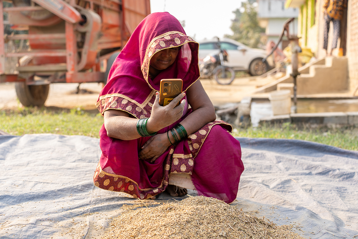 A woman in a bright magenta saree with gold trim sits on a cloth outdoors, holding a smartphone and looking at its screen. In front of her is a small pile of grains. Background shows a tractor, buildings, a parked motorbike, and greenery.