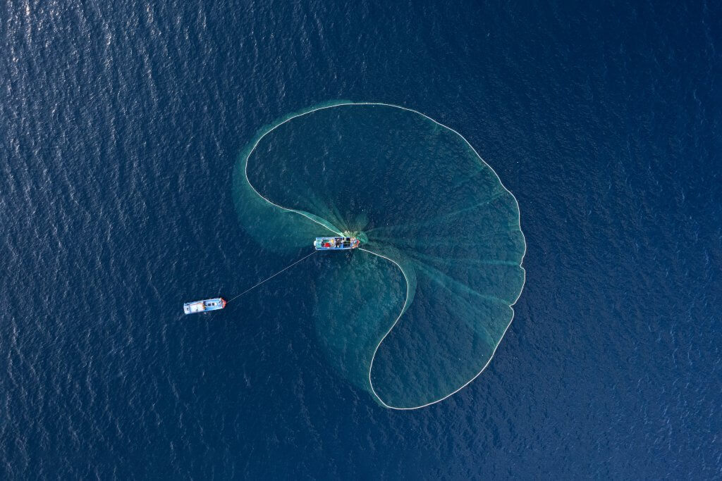 Aerial view of two boats on deep blue water deploying a large, semi-circular fishing net. The net billows outward, creating an impactful, transparent pattern on the water's surface. The boats are connected by a line, and the scene is serene and expansive.