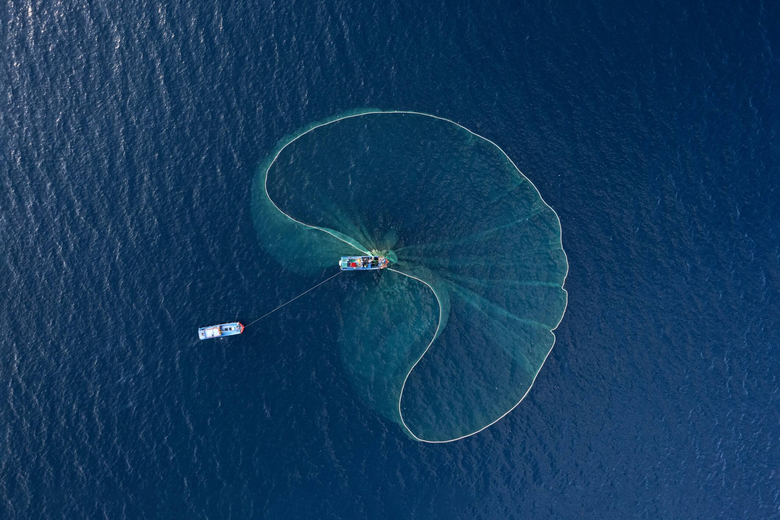 Aerial view of two boats on deep blue water deploying a large, semi-circular fishing net. The net billows outward, creating an impactful, transparent pattern on the water's surface. The boats are connected by a line, and the scene is serene and expansive.