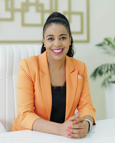 A smiling woman with long dark hair in a high ponytail sits at a white desk. She wears an orange blazer over a black top and has pink nail polish. Behind her are geometric wall art and a green plant. She faces the camera with her hands folded.