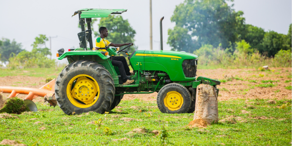 A person drives a green and yellow John Deere tractor across a grassy, slightly muddy field with scattered tree stumps and some farming equipment in the background; trees are visible in the distance under a cloudy sky.