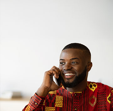 A smiling man wearing a colorful patterned shirt talks on a smartphone. He has a well-groomed short beard and hair, and is sitting indoors against a plain light background. The atmosphere appears bright and cheerful.