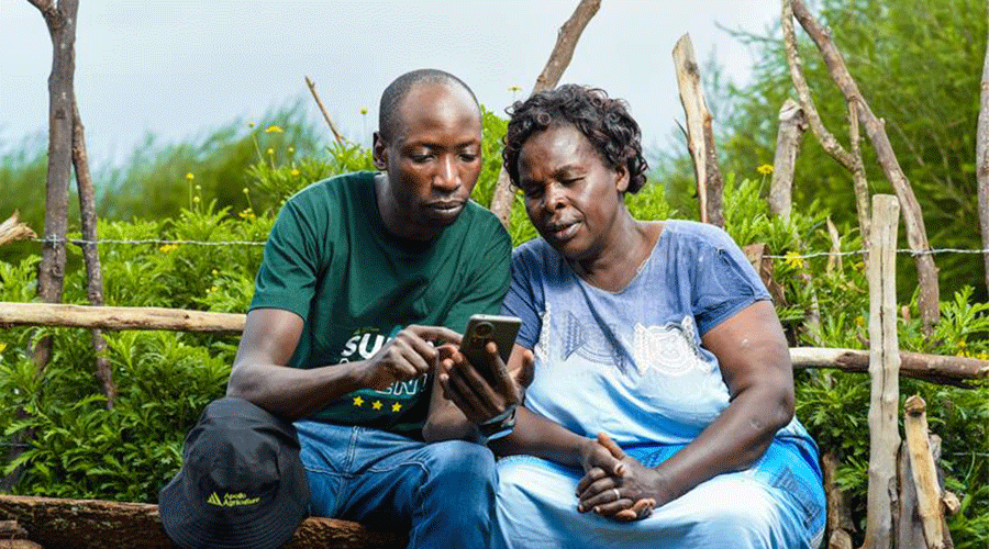 A man in a dark green shirt shows a smartphone to a woman in a blue dress. They sit together on a wooden bench outdoors, surrounded by greenery and rustic wooden fencing. Both are focused on the phone's screen, engaged in a shared activity.