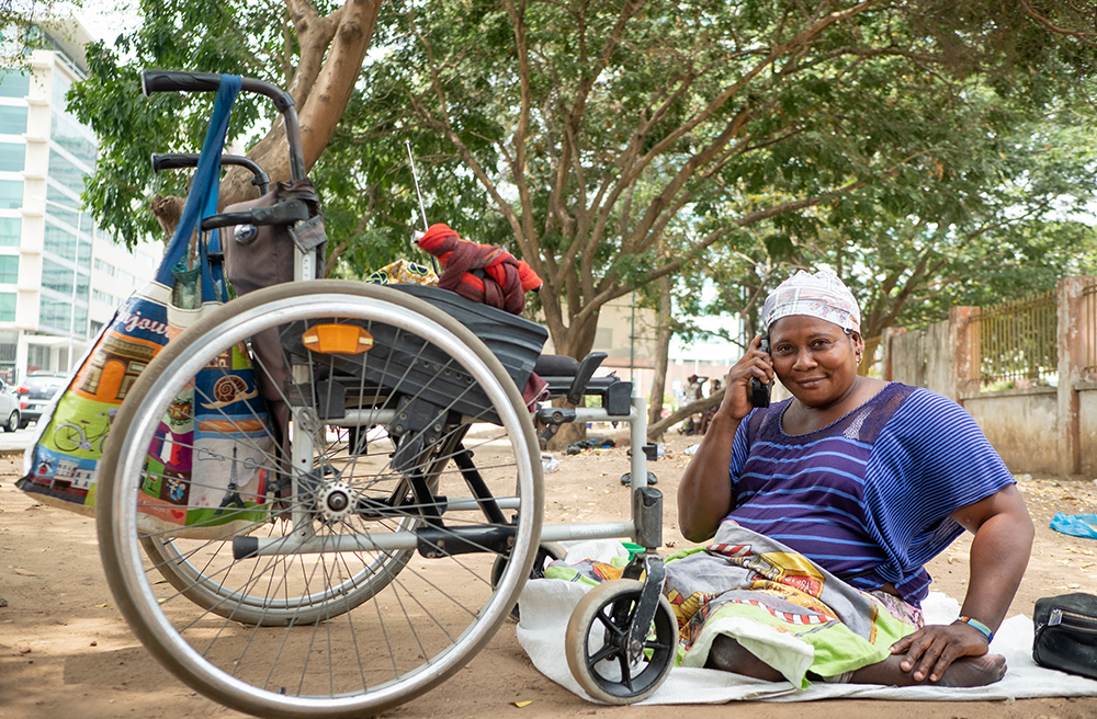 Woman sitting on the ground beside her wheelchair, smiling and talking on a mobile phone under trees in an outdoor setting.