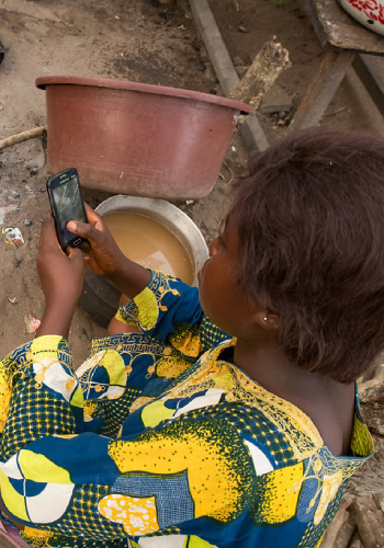 A person with short brown hair, wearing a colorful yellow, blue, and green patterned shirt, sits outdoors holding and looking at a smartphone. In front of them are a brown plastic basin and a round metal bowl filled with brown water.