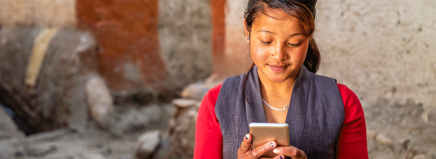 A woman with a nose piercing is focused on her smartphone, wearing a red top and gray vest. Standing outdoors against a rough wall, she seems engaged, epitomizing the spirit of GSMA Mobile for Development in a rustic setting.