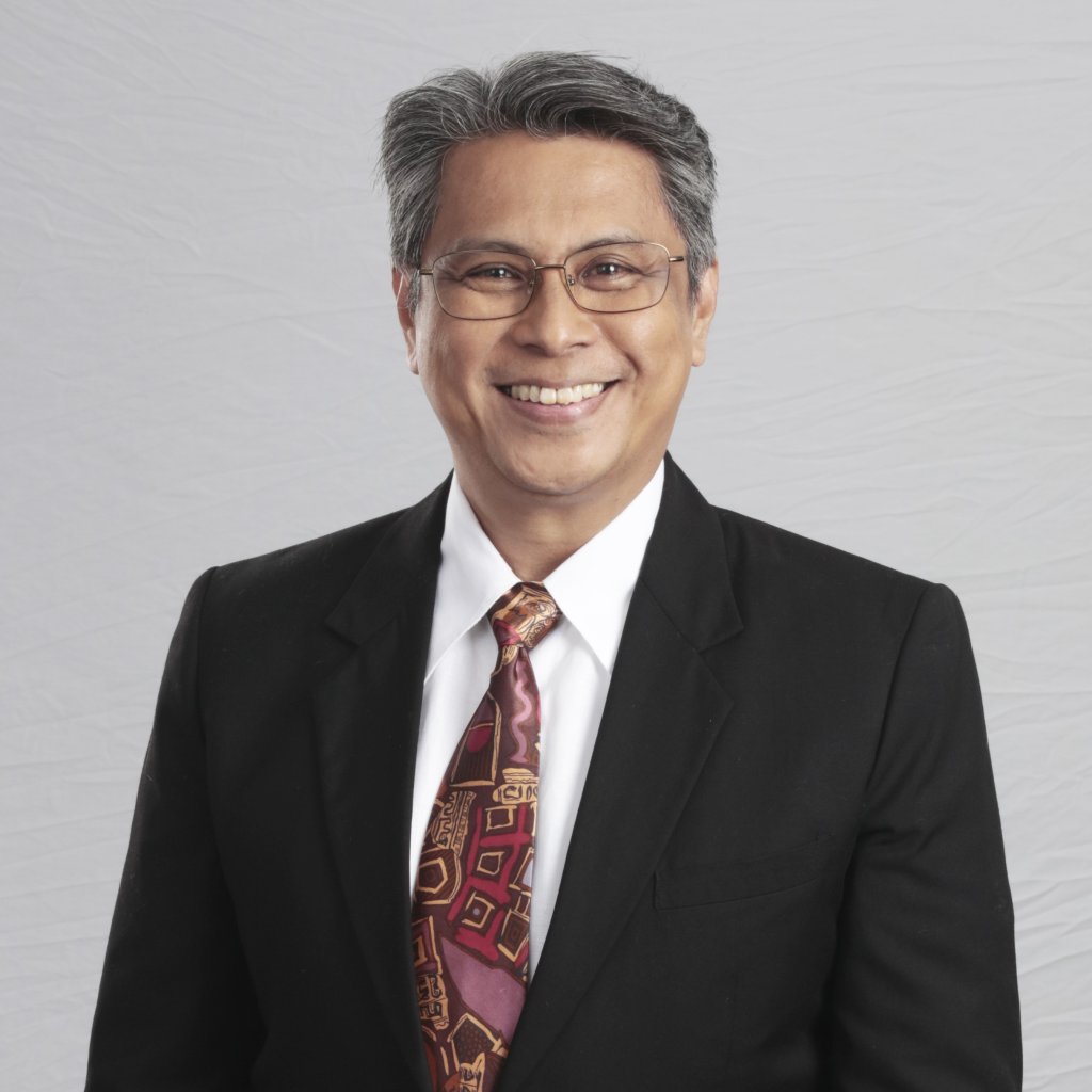 A middle-aged man with short gray hair, glasses, and a friendly smile wears a black suit, white shirt, and a patterned brown and red tie. He stands against a light gray, textured background.