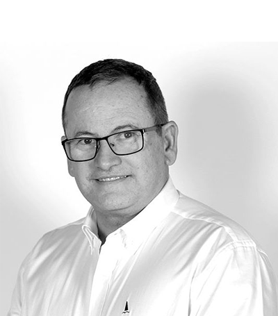 A man with short dark hair, wearing rectangular glasses and a white button-down shirt, smiles slightly while looking at the camera. The background is plain and white. The image is in black and white.