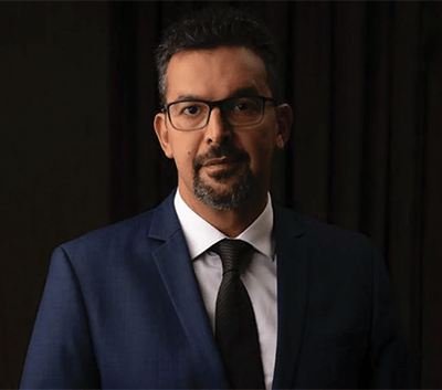 A man with short dark hair, a trimmed beard, and glasses wears a dark blue suit, white shirt, and black tie. He stands against a dark background, looking directly at the camera with a neutral expression.