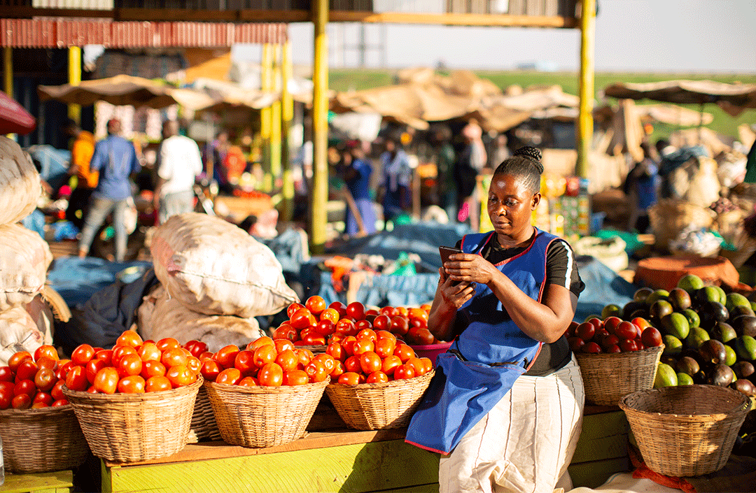 A woman in a blue apron stands behind baskets filled with tomatoes at an open-air market, checking her phone—perhaps for GSMA Mobile for Development updates. Behind her, more vendors display colorful produce among sacks and a bustling, sunlit crowd.