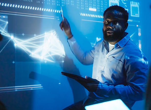 A man in a light blue shirt holds a tablet, gesturing toward a large, futuristic digital screen displaying complex graphs and data under blue lighting. He appears focused and engaged, suggesting an EmergingTech presentation or analysis is in progress.