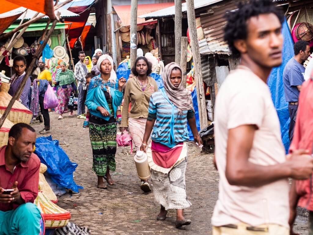 A bustling outdoor market scene unfolds with people walking along the cobblestone street. Vendors make an impact with their vibrant stalls, offering colorful fabrics and goods. In the center, a woman carrying a thermos navigates the lively, crowded atmosphere.