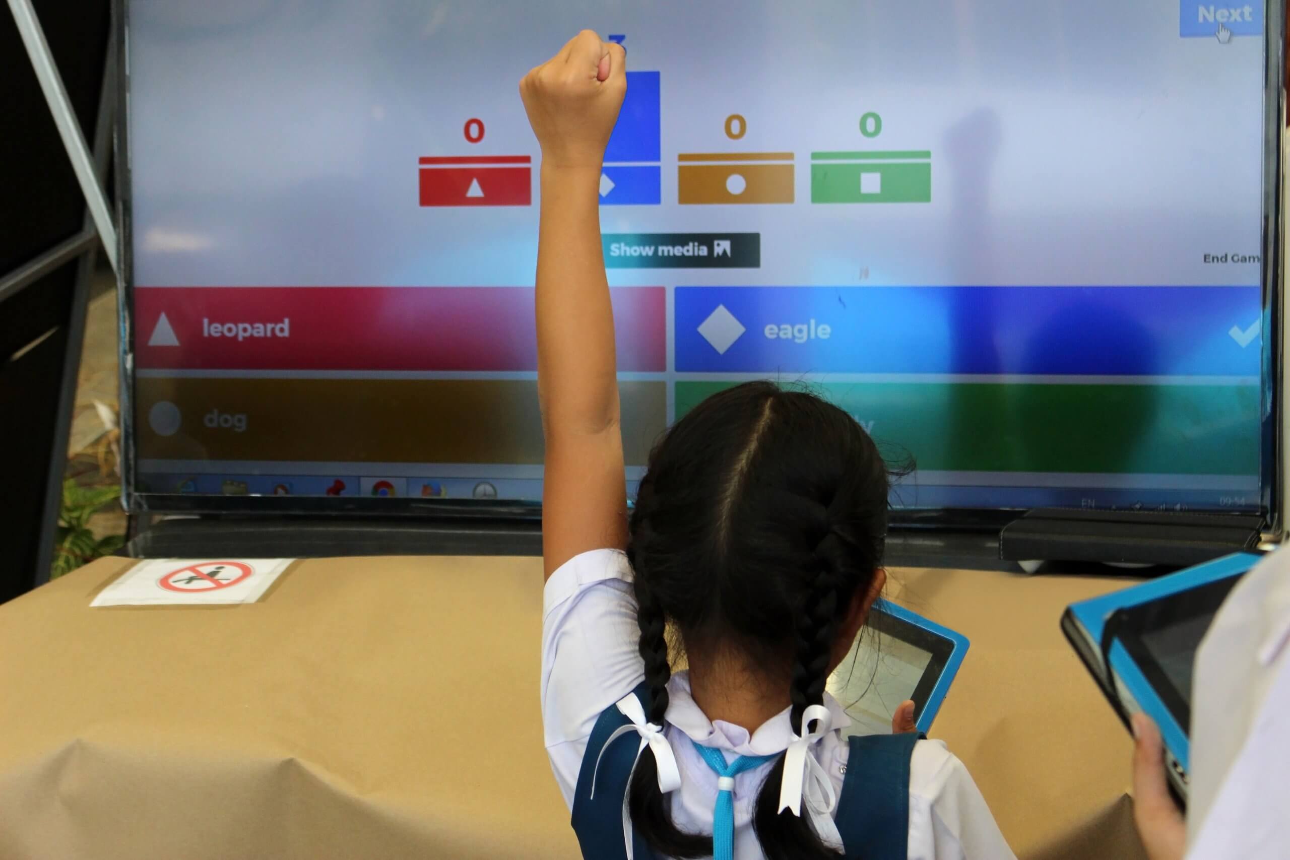 A young girl in a school uniform raises her hand enthusiastically while holding a tablet. She's engaged with an interactive screen displaying a quiz game with color-coded options: red, blue, yellow, and green. The words "leopard" and "eagle" have an impact on her curiosity and learning spirit.