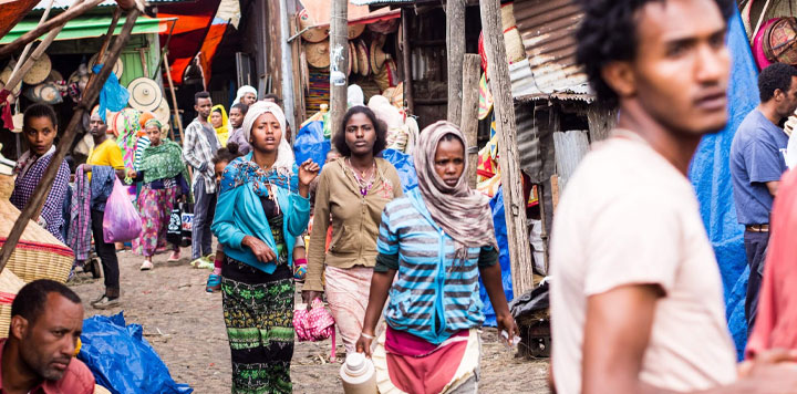 A busy outdoor market scene with people walking and standing. Women in colorful clothes and headscarves carry bags and containers. Market stalls display goods as EmergingTech blends with tradition, under corrugated metal sheets and tarps. The atmosphere is bustling.
