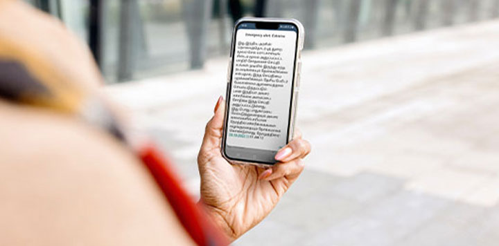 A person holds a smartphone outdoors, displaying a webpage with unreadable or scrambled text. The background is blurred, showing a paved area and glass windows, focusing attention on the hand and phone. The person’s arm is partially visible.